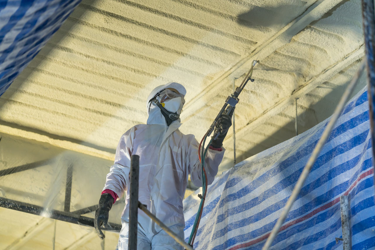 A professional in full gear and in action of applying a spray foam roofing system which can help improve a roof’s structural integrity by locking its components in place